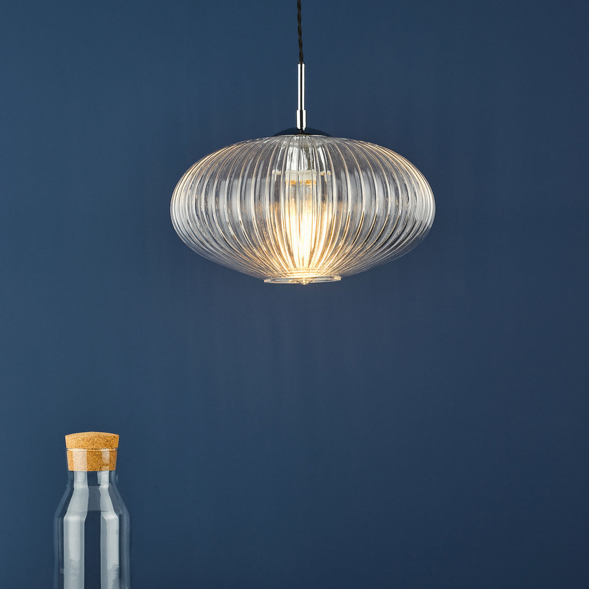 Ribbed glass pendant light with a warm glow, hanging against a dark blue wall. A glass bottle with a cork top is at the bottom left.