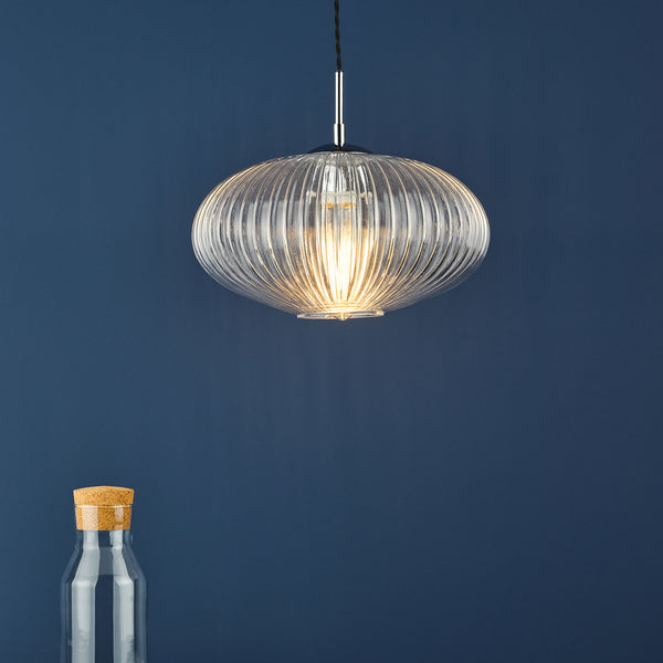 Ribbed glass pendant light with a warm glow, hanging against a dark blue wall. A glass bottle with a cork top is at the bottom left.