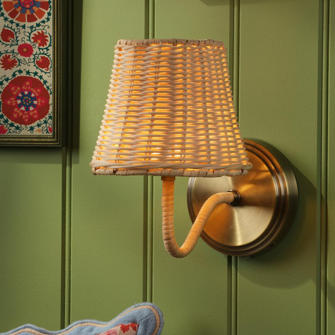 Woven rattan wall sconce with brass backplate mounted on a green paneled wall