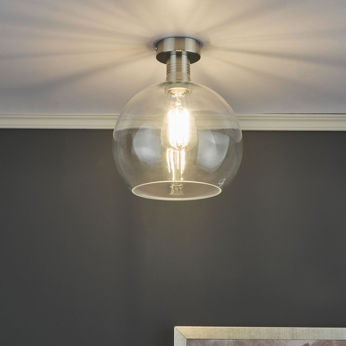 A clear glass globe ceiling light with a brushed nickel base, illuminating a dark gray wall.
