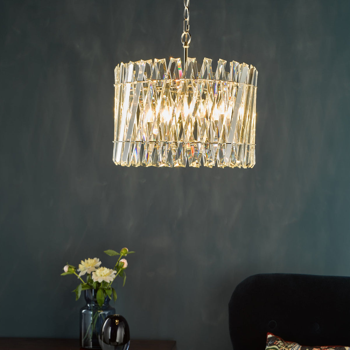 Rectangular crystal chandelier with a gold chain, dark blue wall, and flowers in a vase