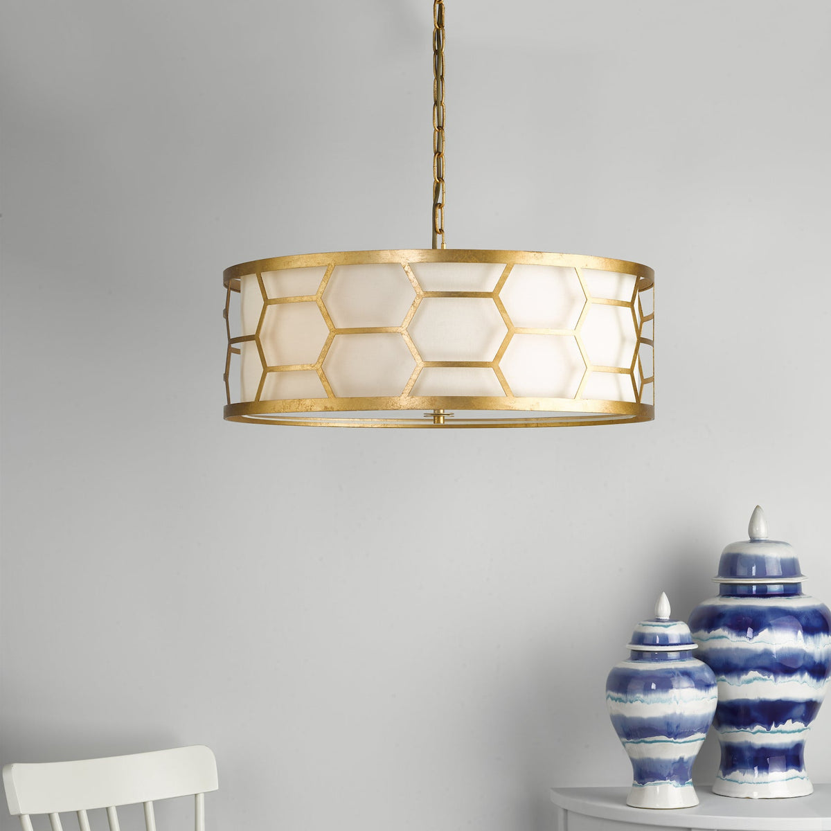 Gold chandelier with a honeycomb pattern and a white drum shade, hanging above two blue and white decorative jars