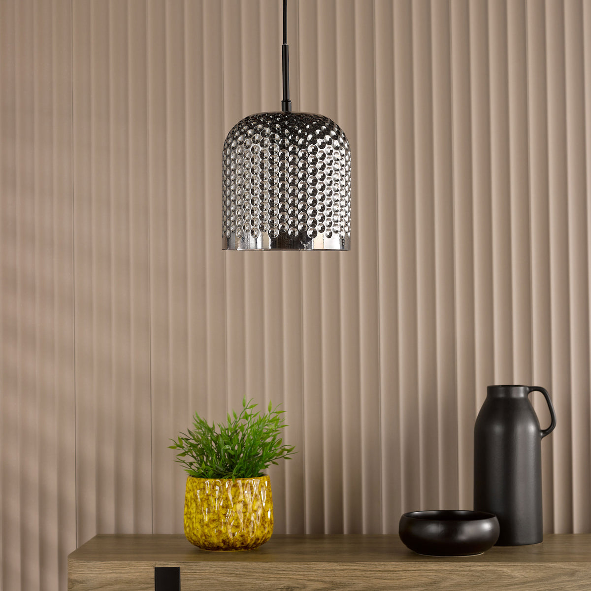 Silver pendant light with textured surface, green plant in yellow pot, black vase and bowl on wooden surface