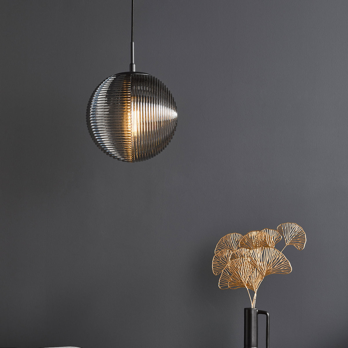 A modern ribbed glass pendant light hangs above a black vase with gold decorative leaves.