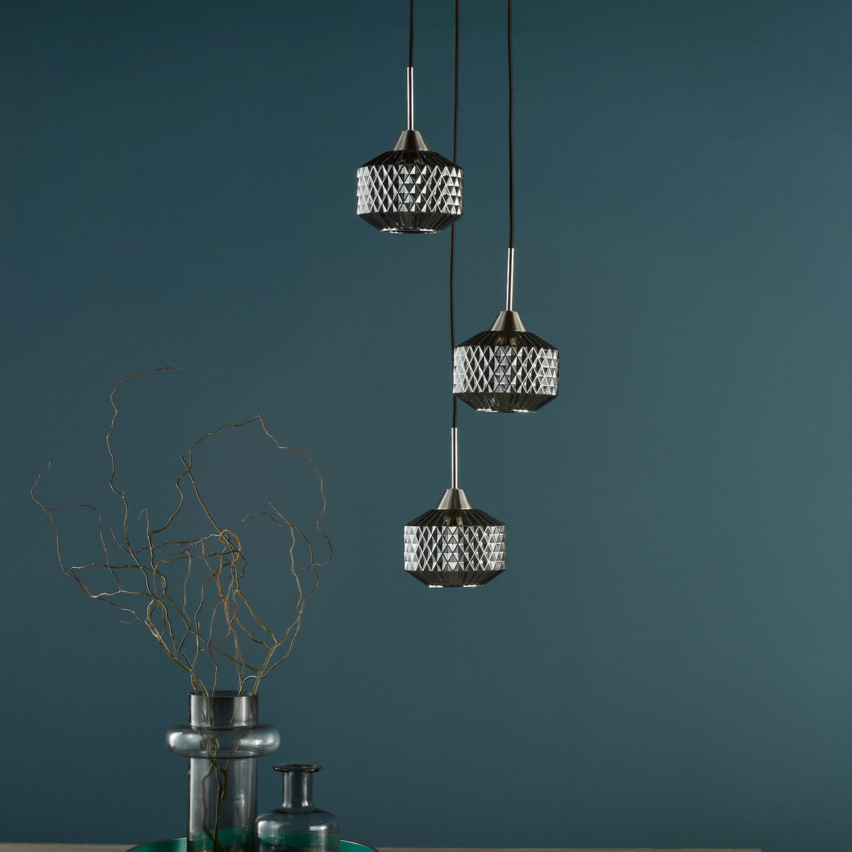 Three pendant lights with faceted glass shades hang against a dark teal wall, with a vase of branches below