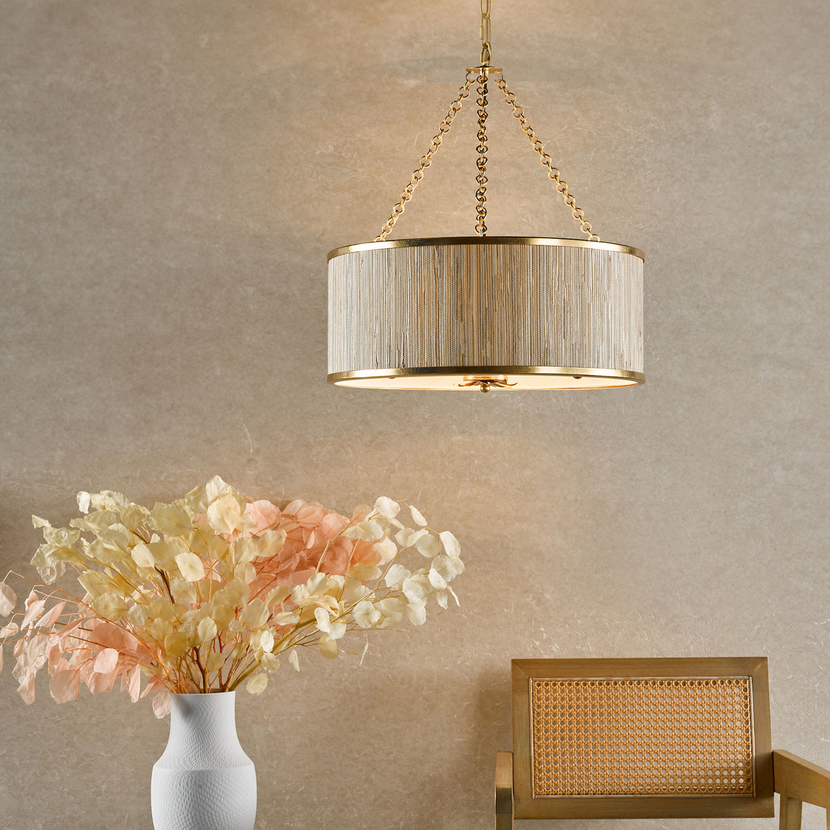 A gold chandelier with a textured drum shade hangs above a white vase with dried flowers and a cane chair.