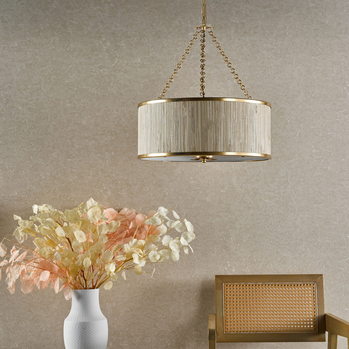 A gold chandelier with a woven shade hangs above a white vase with dried flowers and a wooden chair.