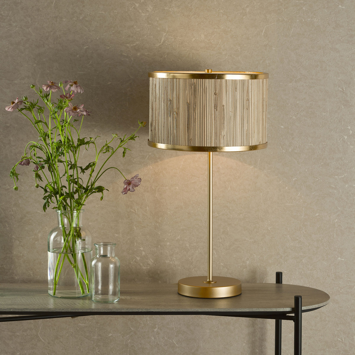 A gold table lamp with a woven shade next to a vase of pink flowers on a wooden console table.