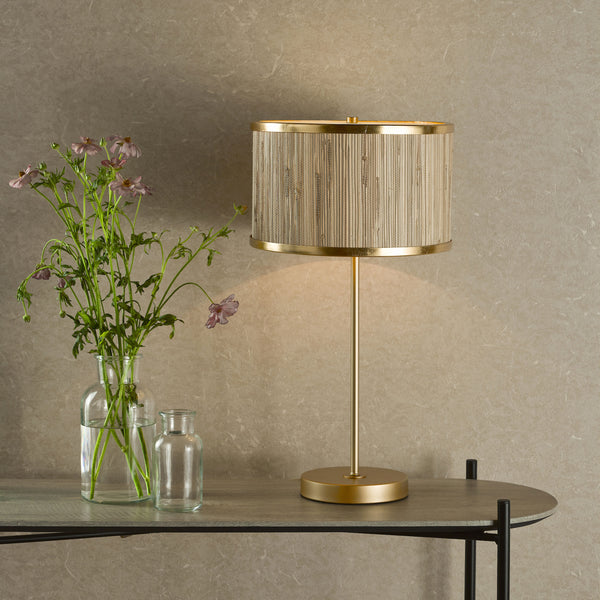 A gold table lamp with a woven shade next to a vase of pink flowers on a wooden console table.