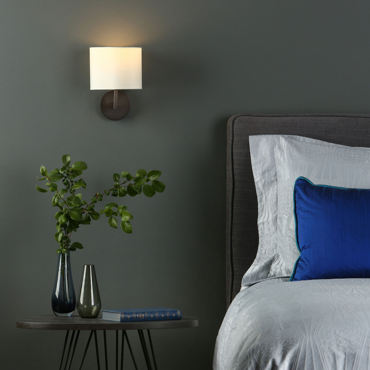 A bedroom with a dark gray wall, a wall sconce, a nightstand with two vases and a book, and a bed.