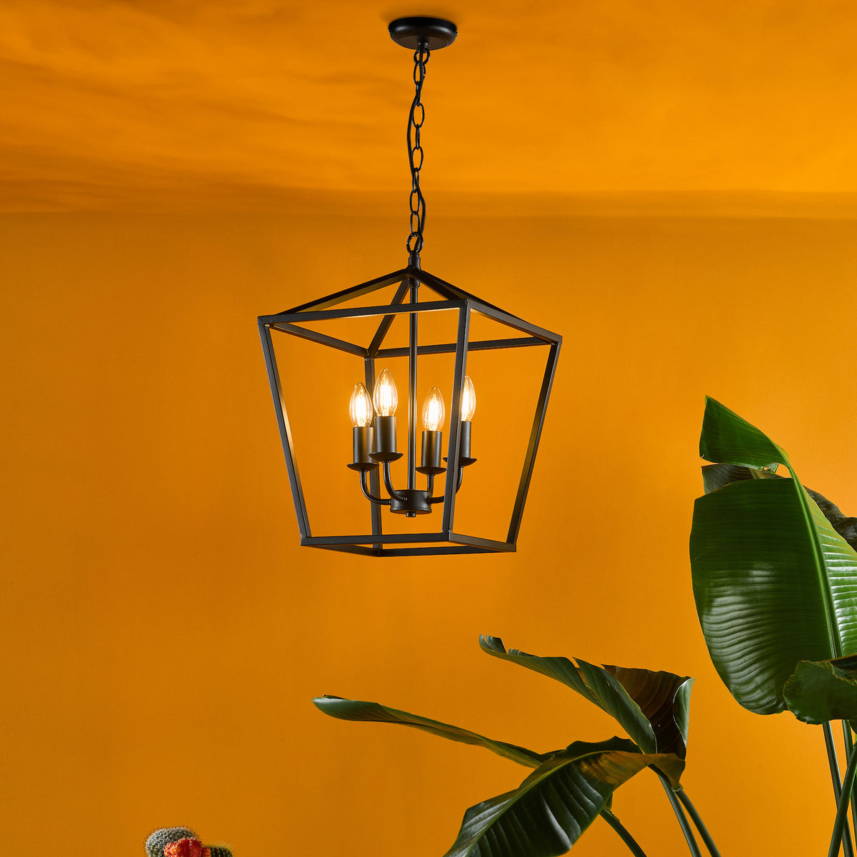 Black metal geometric chandelier with four candle-style bulbs, against a vibrant orange wall with green plant leaves.