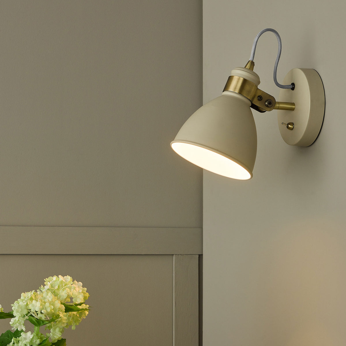 Cream wall light with brass accents and a flexible arm, illuminating a wall and light green flowers