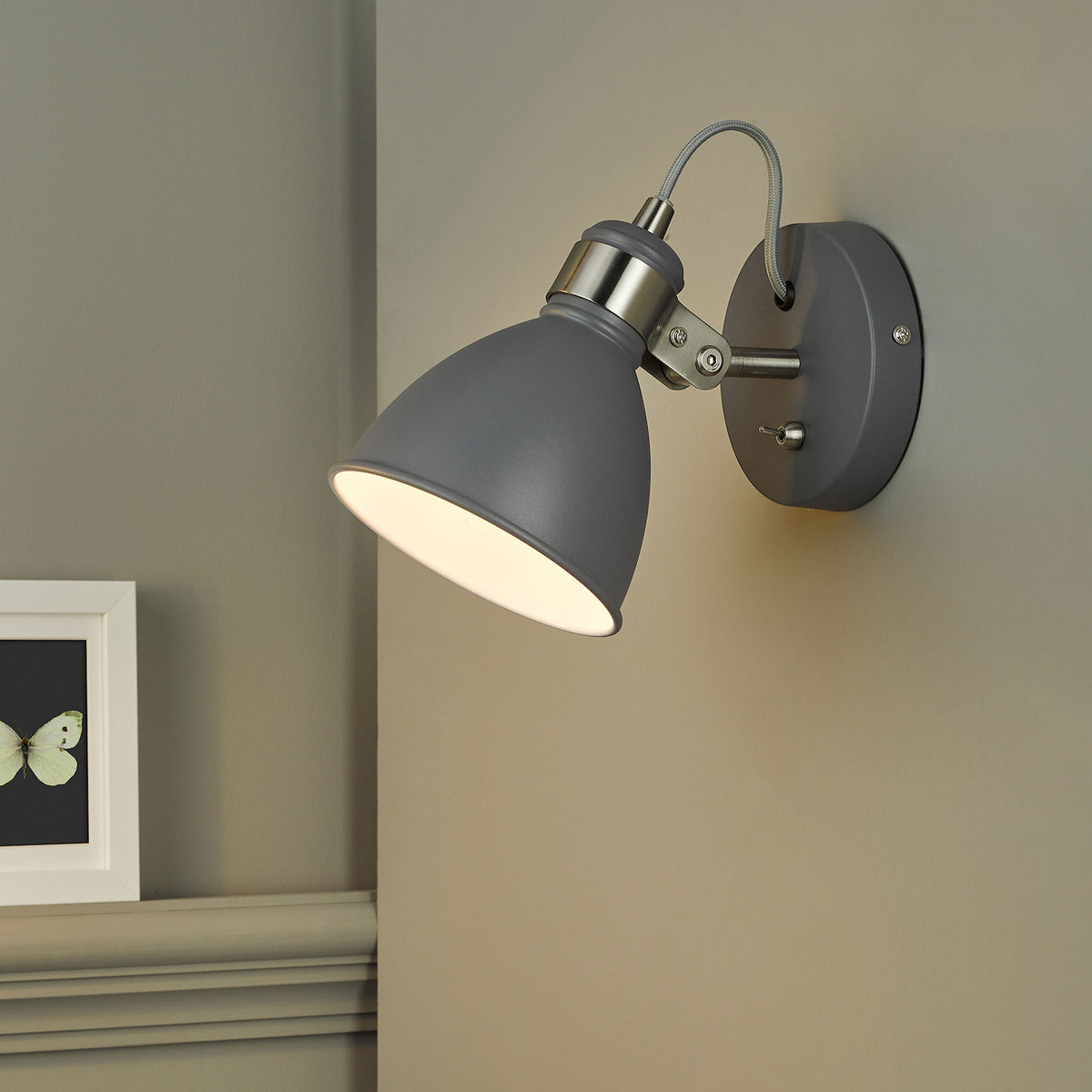 Grey wall light with a brushed steel arm and a toggle switch, illuminating a wall and a framed butterfly print