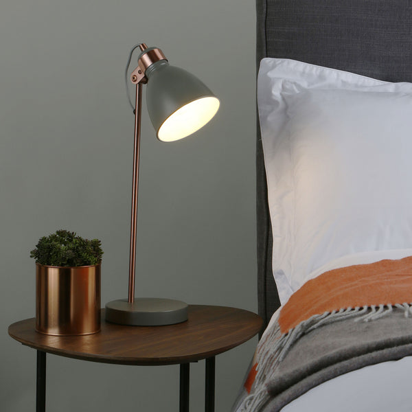 A gray and copper desk lamp illuminates a bedside table with a small plant next to a bed with white sheets and an orange and gray blanket.