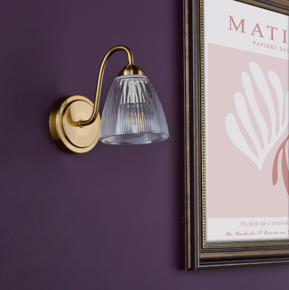 Brass wall light with a fluted glass shade on a purple wall next to a framed poster
