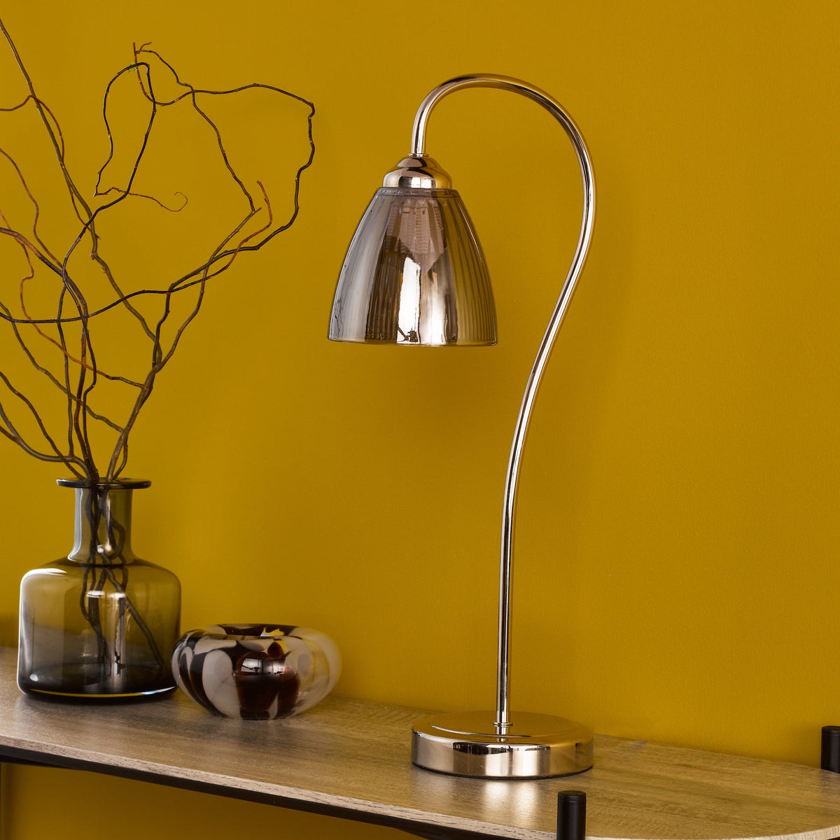 Polished chrome table lamp with a curved stem and a ribbed glass shade on a wooden shelf