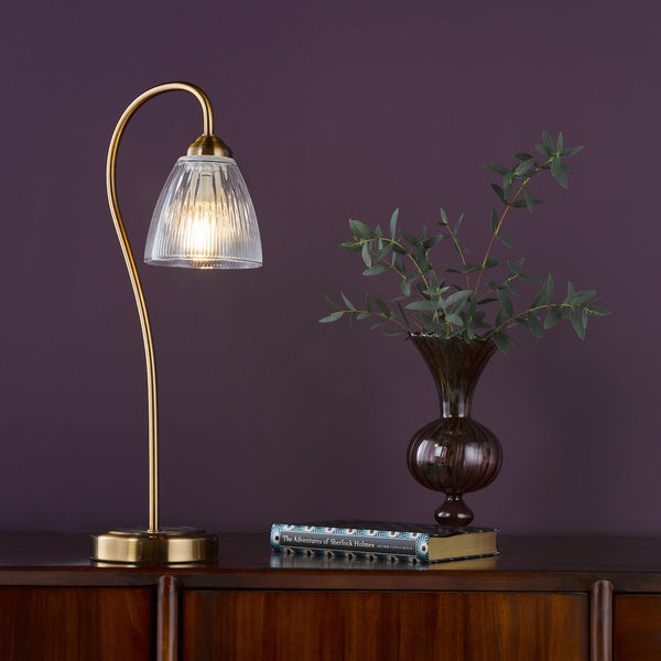 Brass table lamp with a fluted glass shade on a wooden cabinet next to a vase with foliage