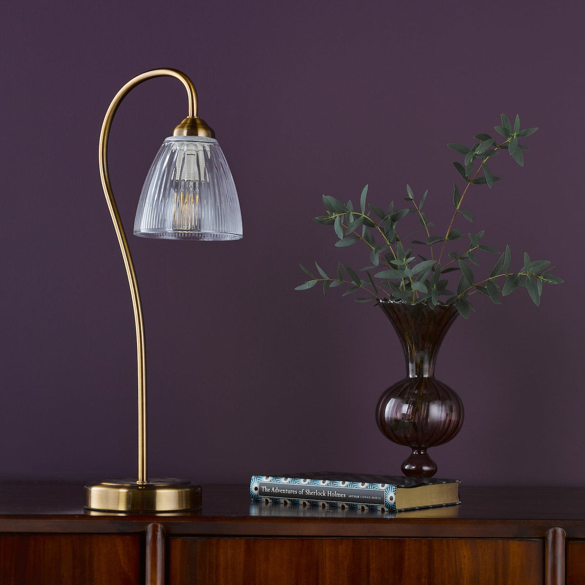 Brass table lamp with a fluted glass shade, a dark vase with foliage, and a book on a wooden surface