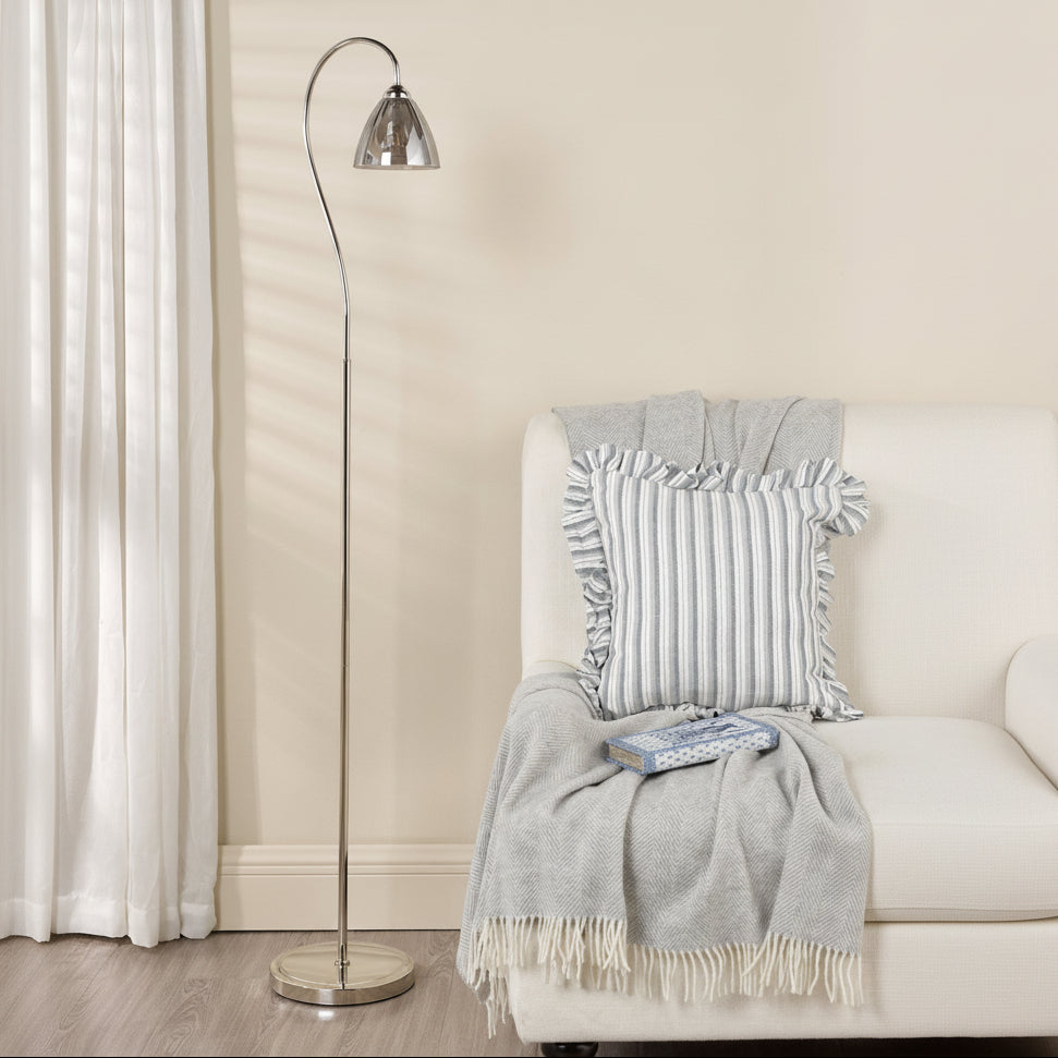 Silver floor lamp with a curved neck and a clear glass shade next to a white armchair with a grey throw