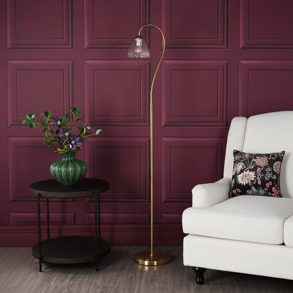 A gold floor lamp with a clear glass shade next to a white armchair with a floral cushion
