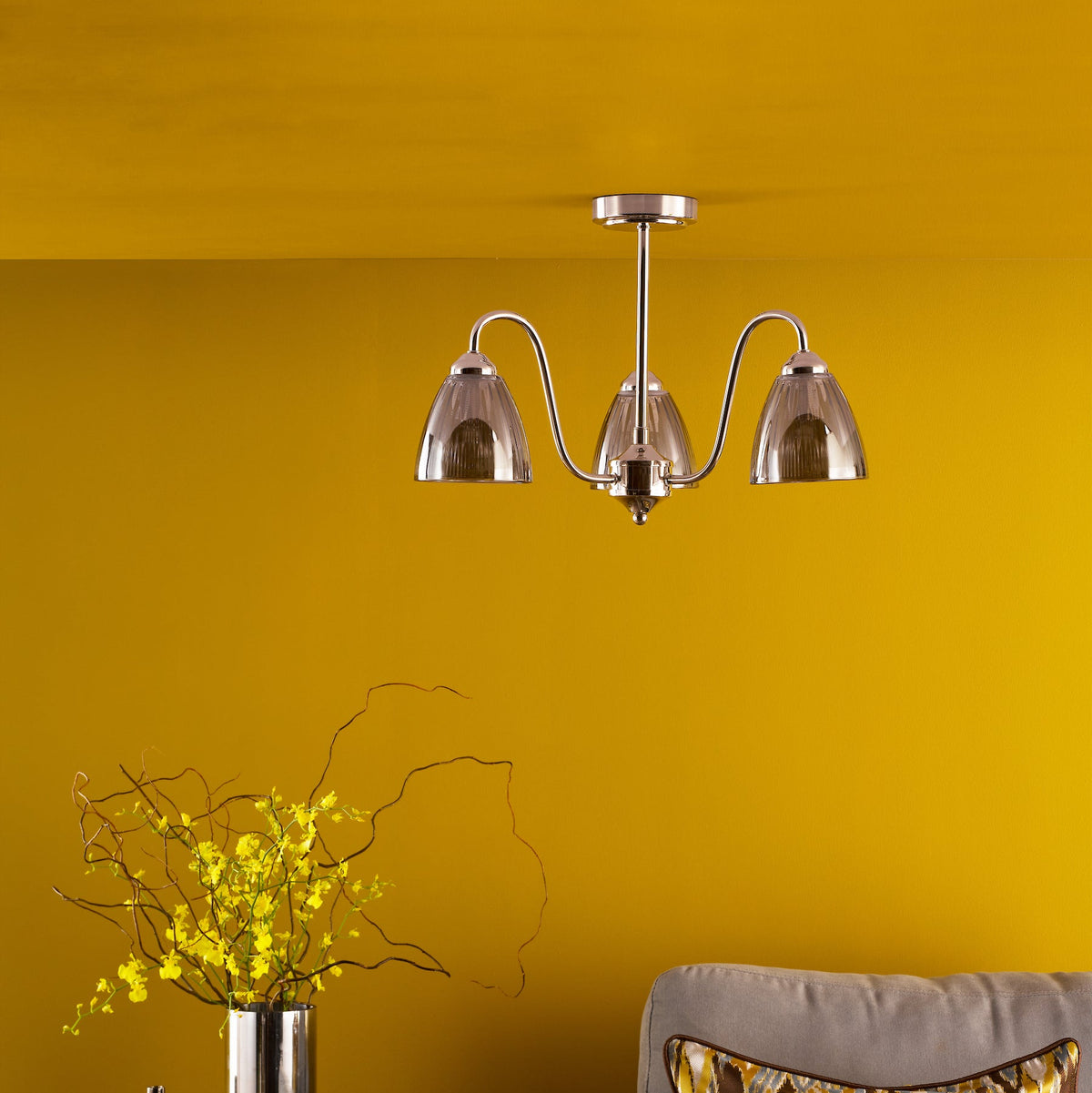 Three light chandelier with clear glass shades on a yellow wall above a grey sofa and yellow flowers