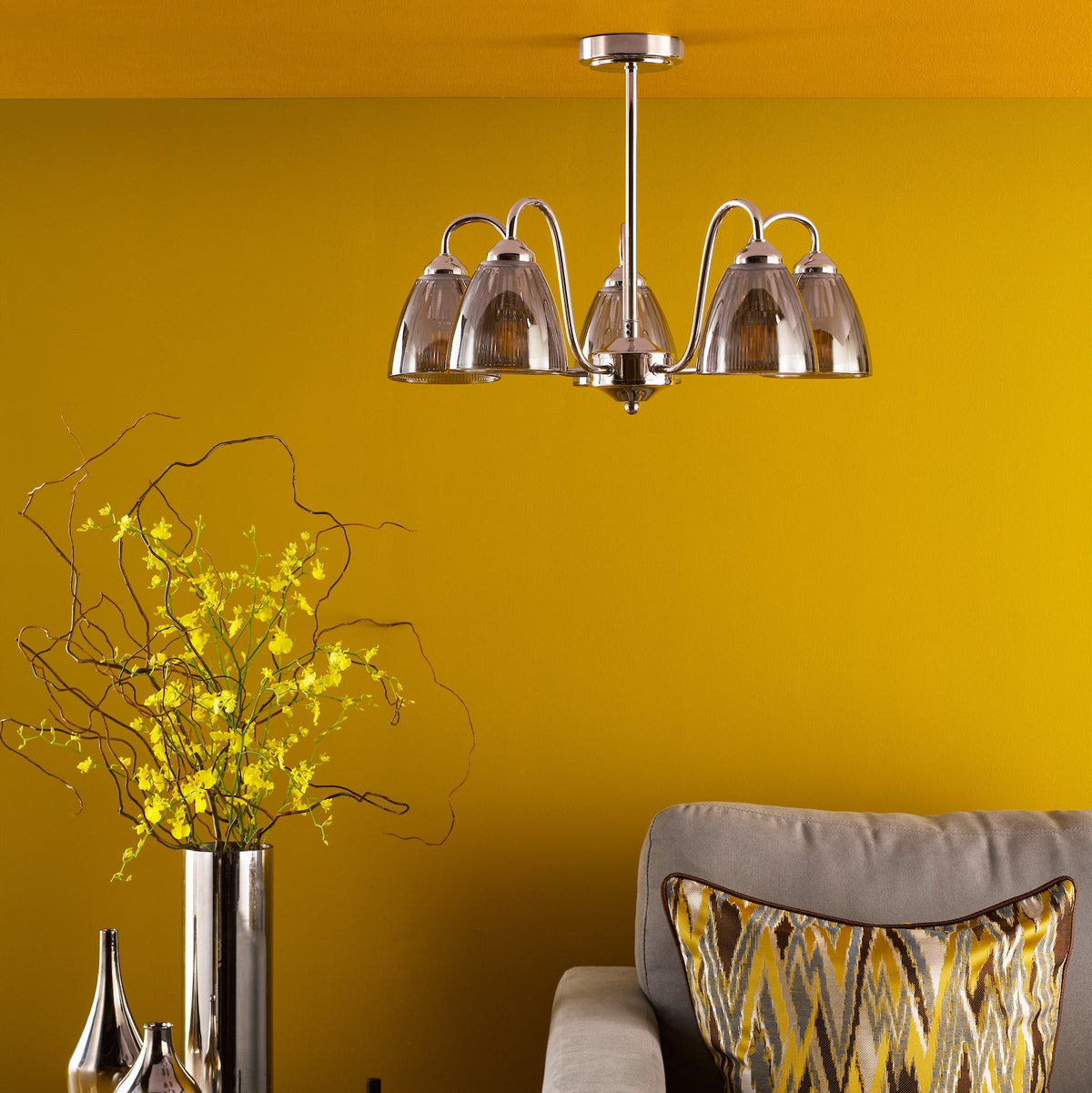 Polished chrome chandelier with four ribbed glass shades, above a vase of yellow flowers and a grey sofa