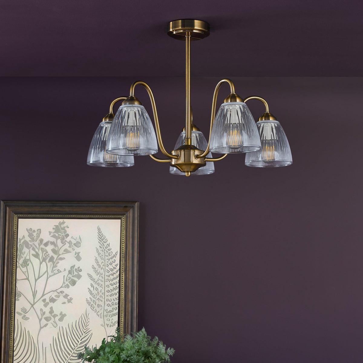 Brass chandelier with five ribbed glass shades, illuminating a dark purple wall with framed botanical art