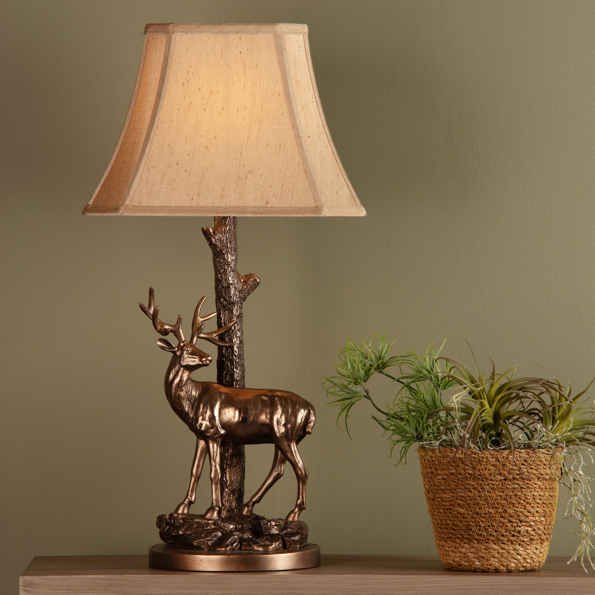 Bronze deer lamp with a beige shade next to a woven pot with green plants.