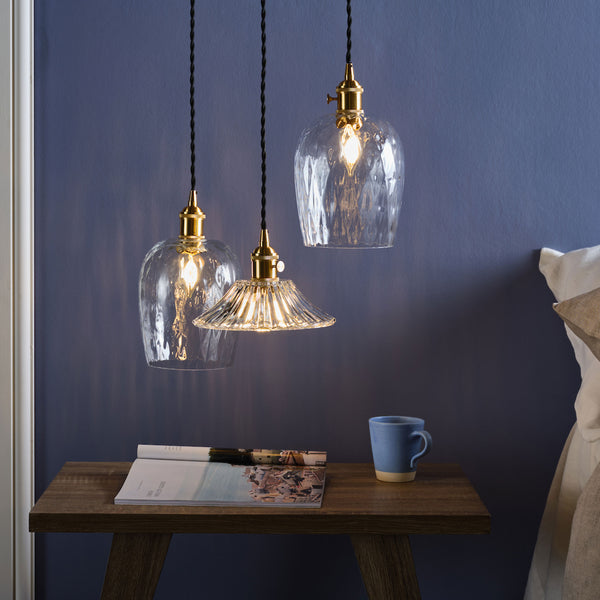 Three clear glass pendant lights with brass fittings hang above a wooden bedside table with a magazine and mug.