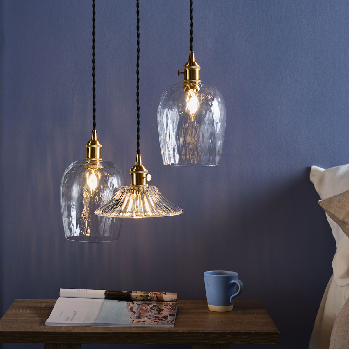 Three pendant lights with clear glass shades hang above a wooden bedside table with a magazine and mug.