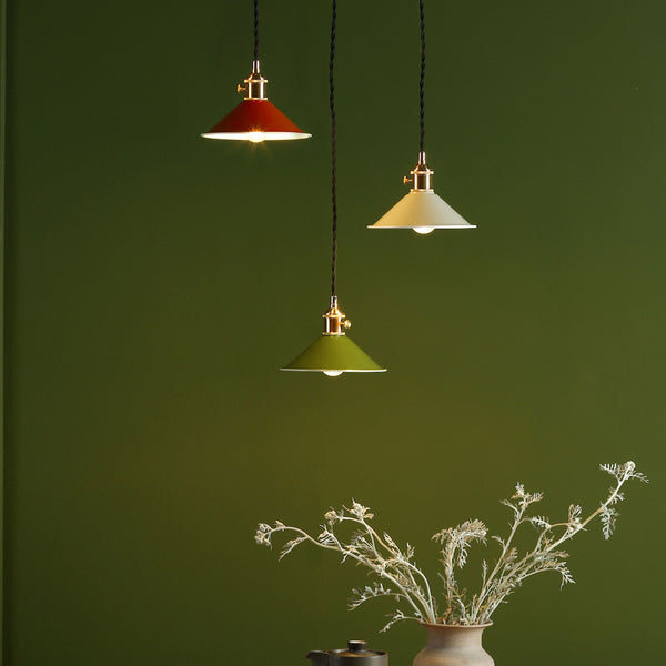 Three pendant lights in red, cream, and green hang against a dark green wall. Dried white foliage in a vase below.