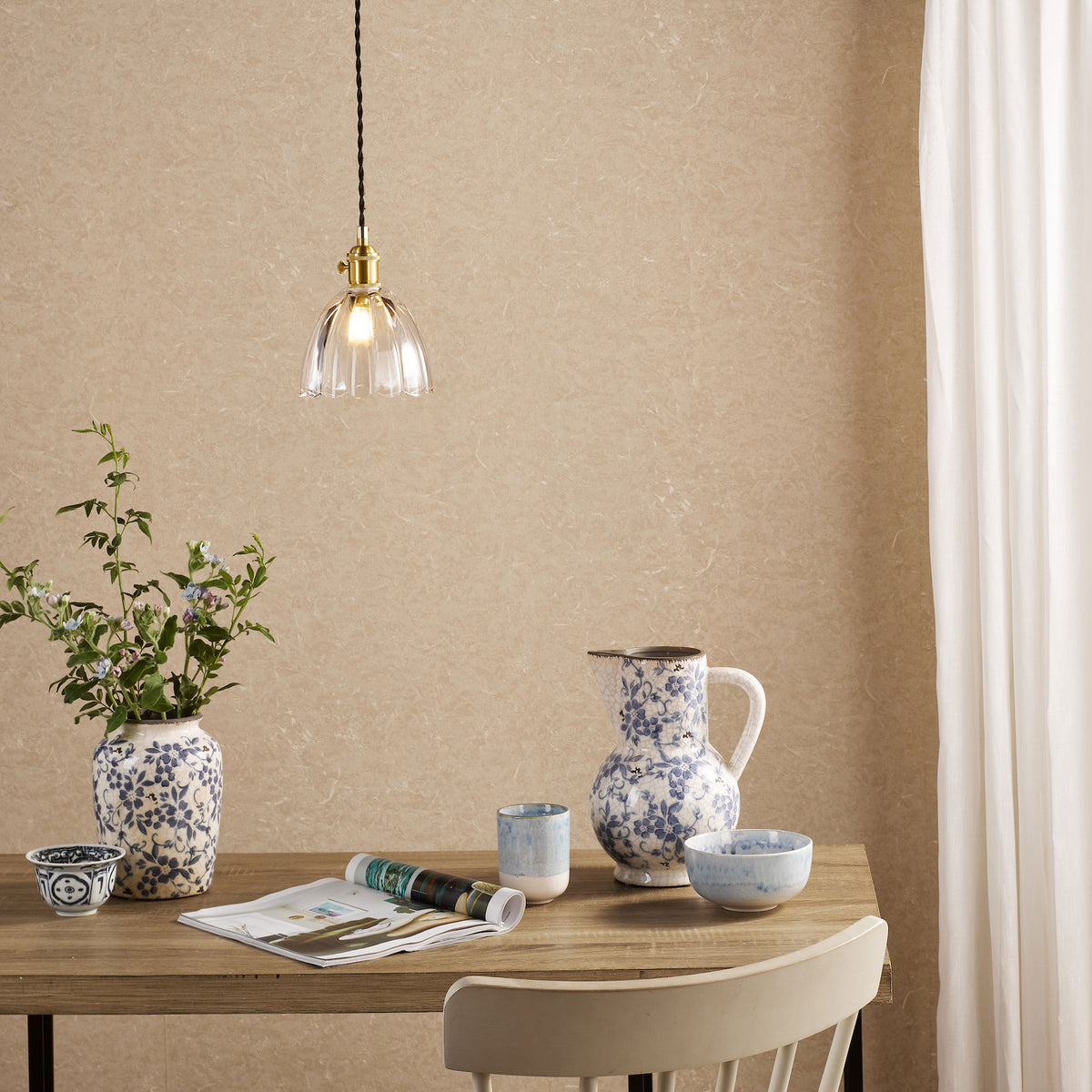 A dining table with a vase of flowers, a pitcher, and bowls, under a hanging glass lamp.