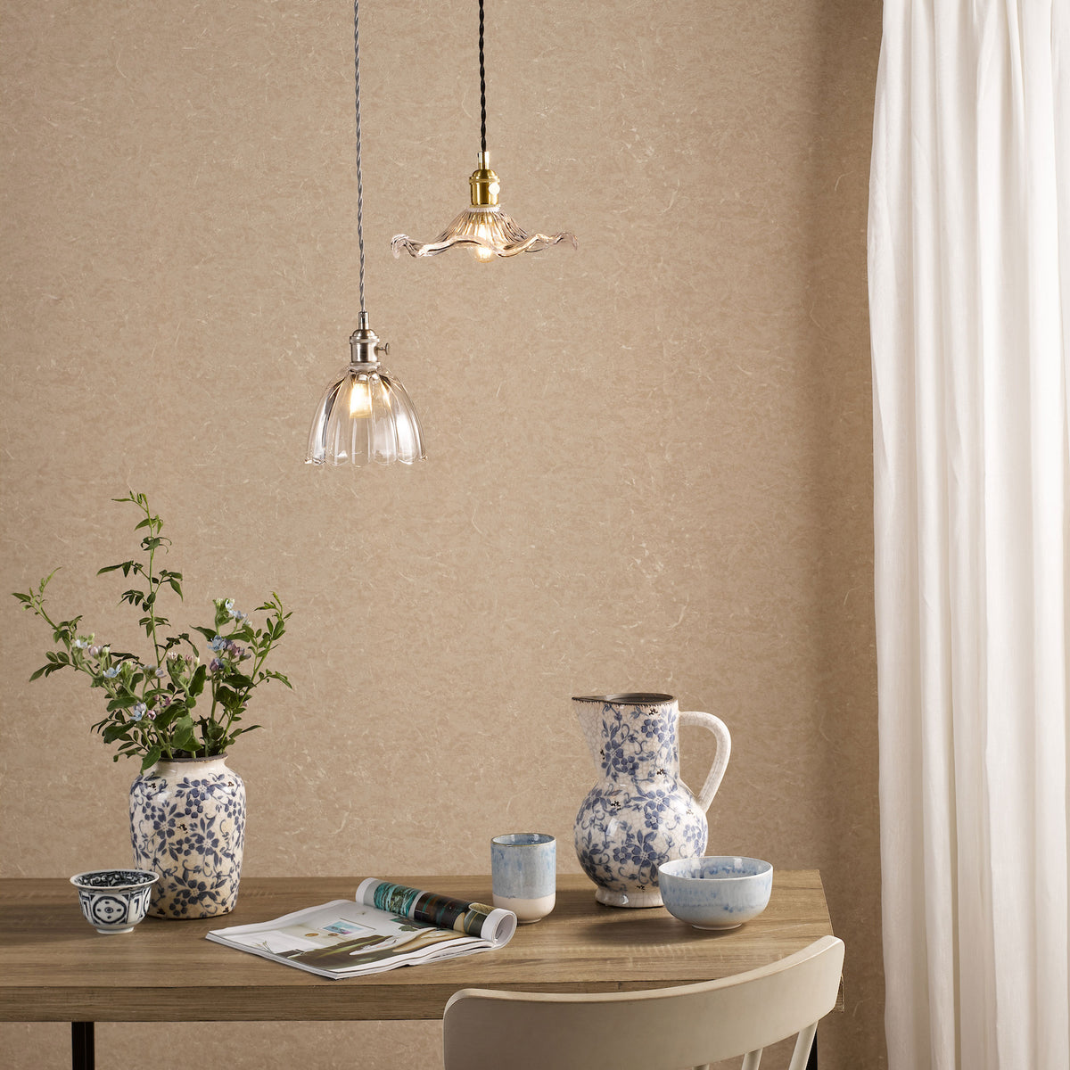 Two glass pendant lights hang above a wooden table with blue and white patterned ceramics and a magazine.