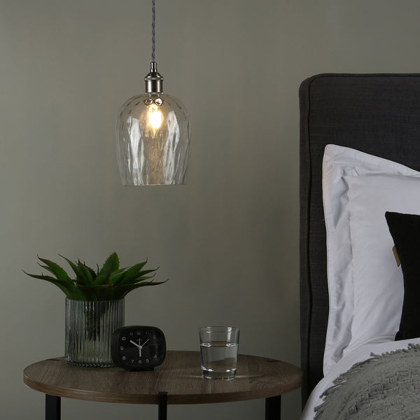 A clear glass pendant light hangs above a bedside table with a plant, clock, and glass of water.