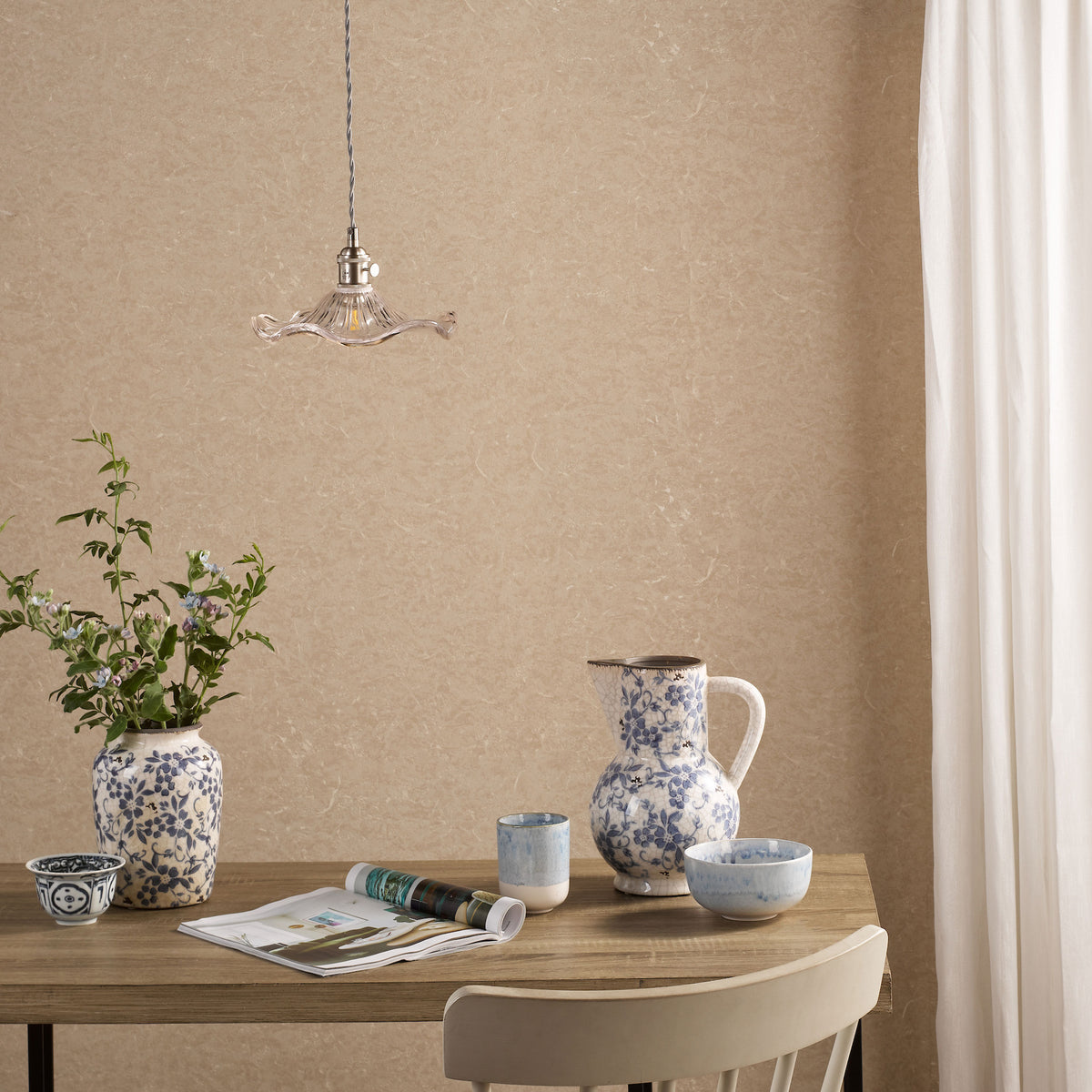 A dining table with a vase of flowers, a pitcher, and bowls, all with blue and white patterns. A magazine lies open on the table. A light fixture hangs above.