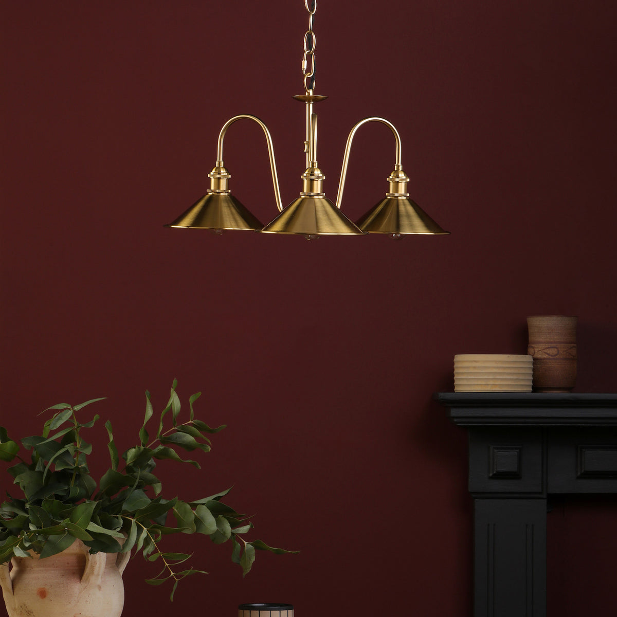 A brass three-arm chandelier hangs from a chain against a deep red wall, with plants and pottery below.