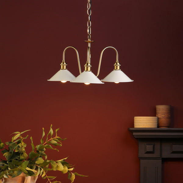 Three-light chandelier with white shades and brass accents, hanging against a deep red wall. Greenery in foreground.
