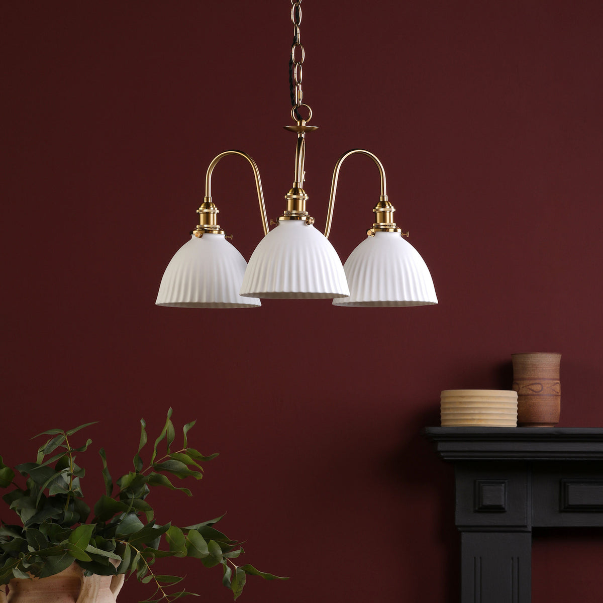 Three-light brass chandelier with ribbed white shades against a deep red wall. A plant and vases are on a mantelpiece.