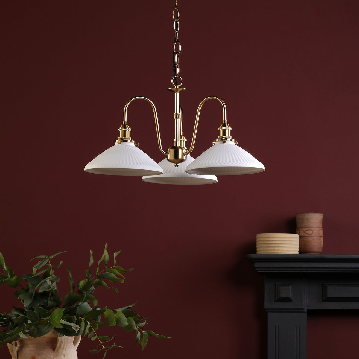 A gold chandelier with three white, fluted shades hangs against a deep red wall. A black mantelpiece is to the right.