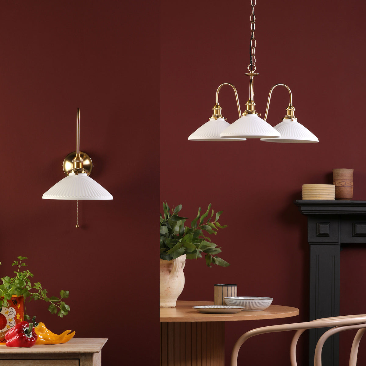 A brass wall sconce and a three-light chandelier with pleated white shades against a deep red wall.