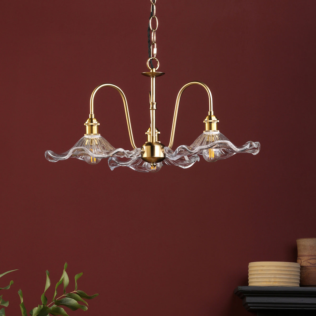 A brass chandelier with three wavy glass shades hangs against a deep red wall. A plant and pottery are below.