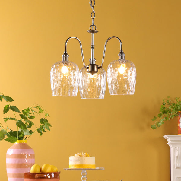 A three-light chandelier with clear, textured glass shades hangs against a yellow wall. A cake and lemons are on the table.
