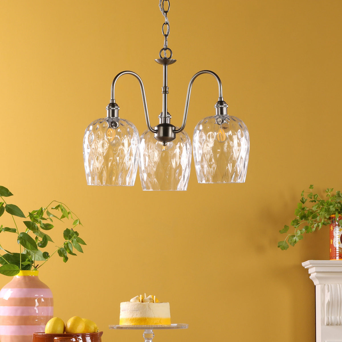 Three-light chandelier with clear, textured glass shades against a yellow wall. A cake, lemons, and plants are below.