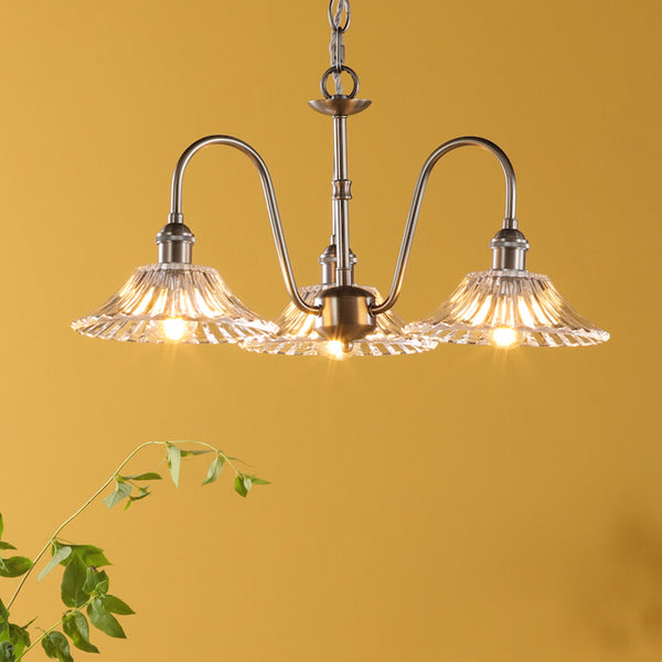 A three-arm chandelier with clear, fluted glass shades and a brushed brass finish, against a yellow wall.