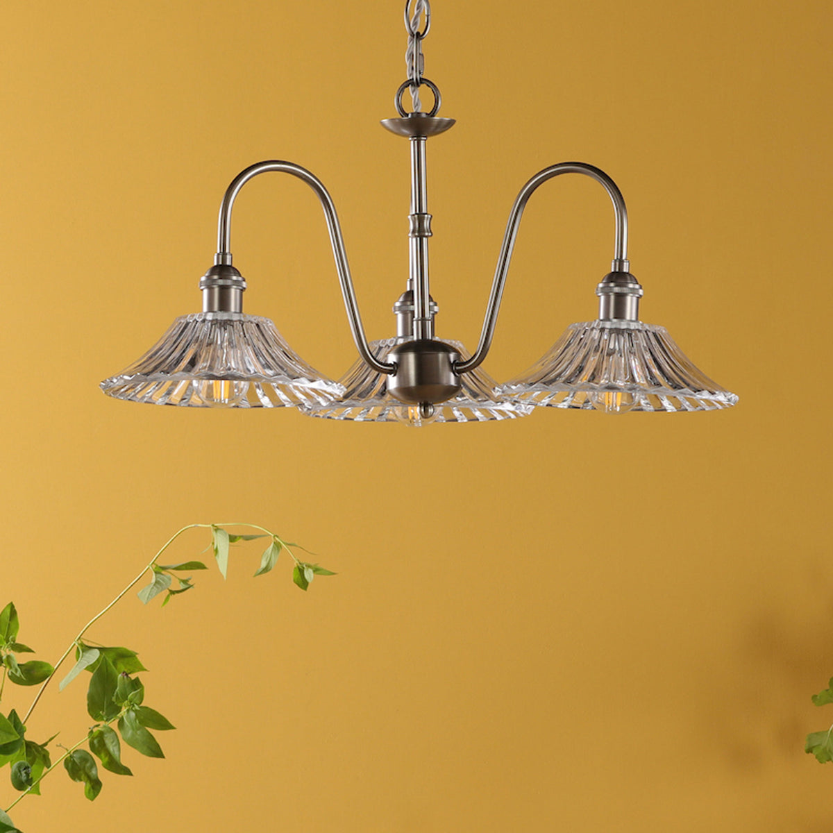 A three-arm chandelier with clear, fluted glass shades and a brushed nickel finish, against a mustard yellow wall.