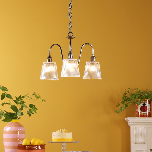 Three-light chandelier with ribbed glass shades, hanging against a yellow wall. A cake, lemons, and plants are below.