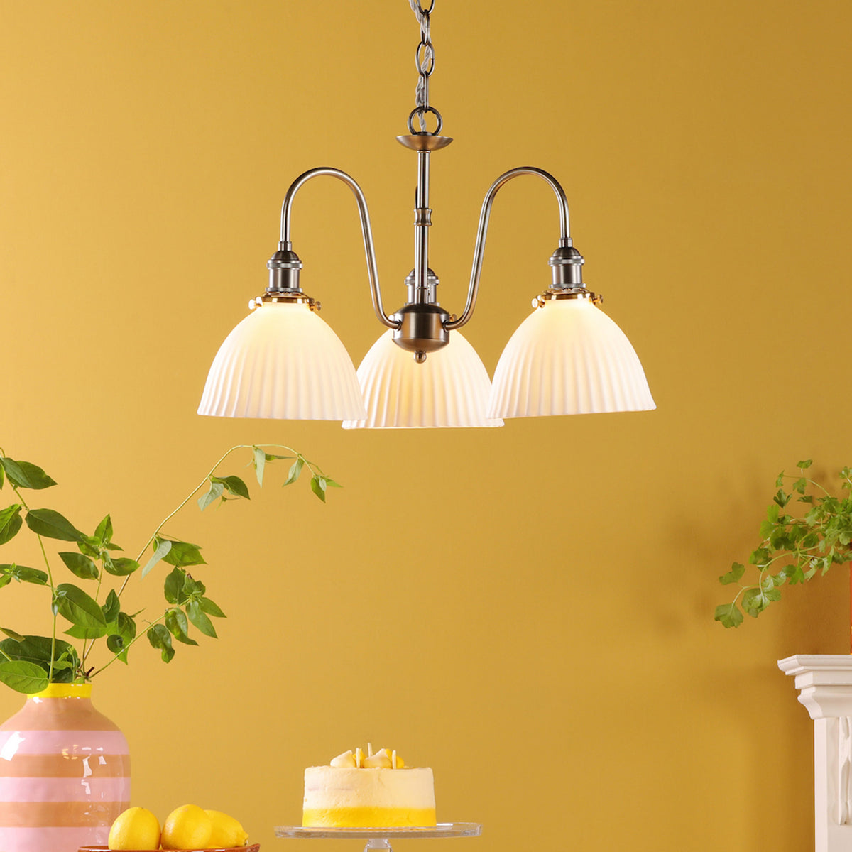 A three-light chandelier with pleated glass shades hangs against a yellow wall, above a cake and lemons.