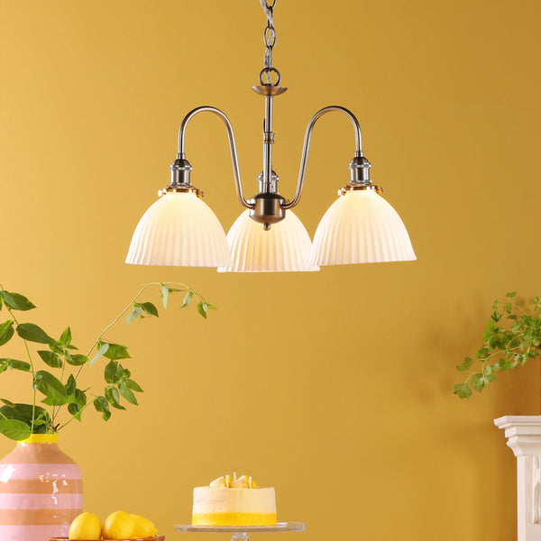 A three-light chandelier with pleated glass shades hangs against a yellow wall, above a cake and lemons.