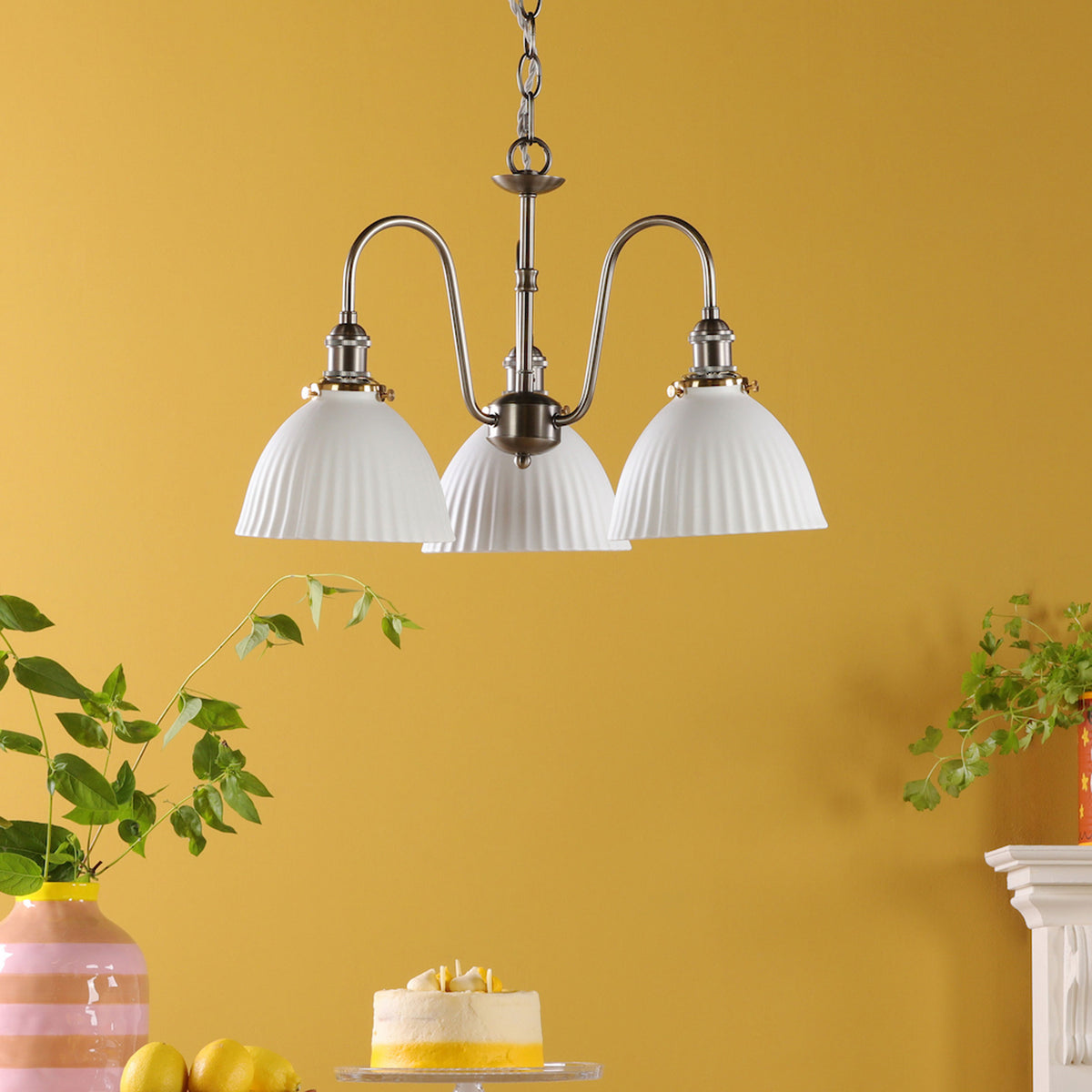 A three-light chandelier with white fluted shades hangs against a yellow wall, with a cake and lemons below.