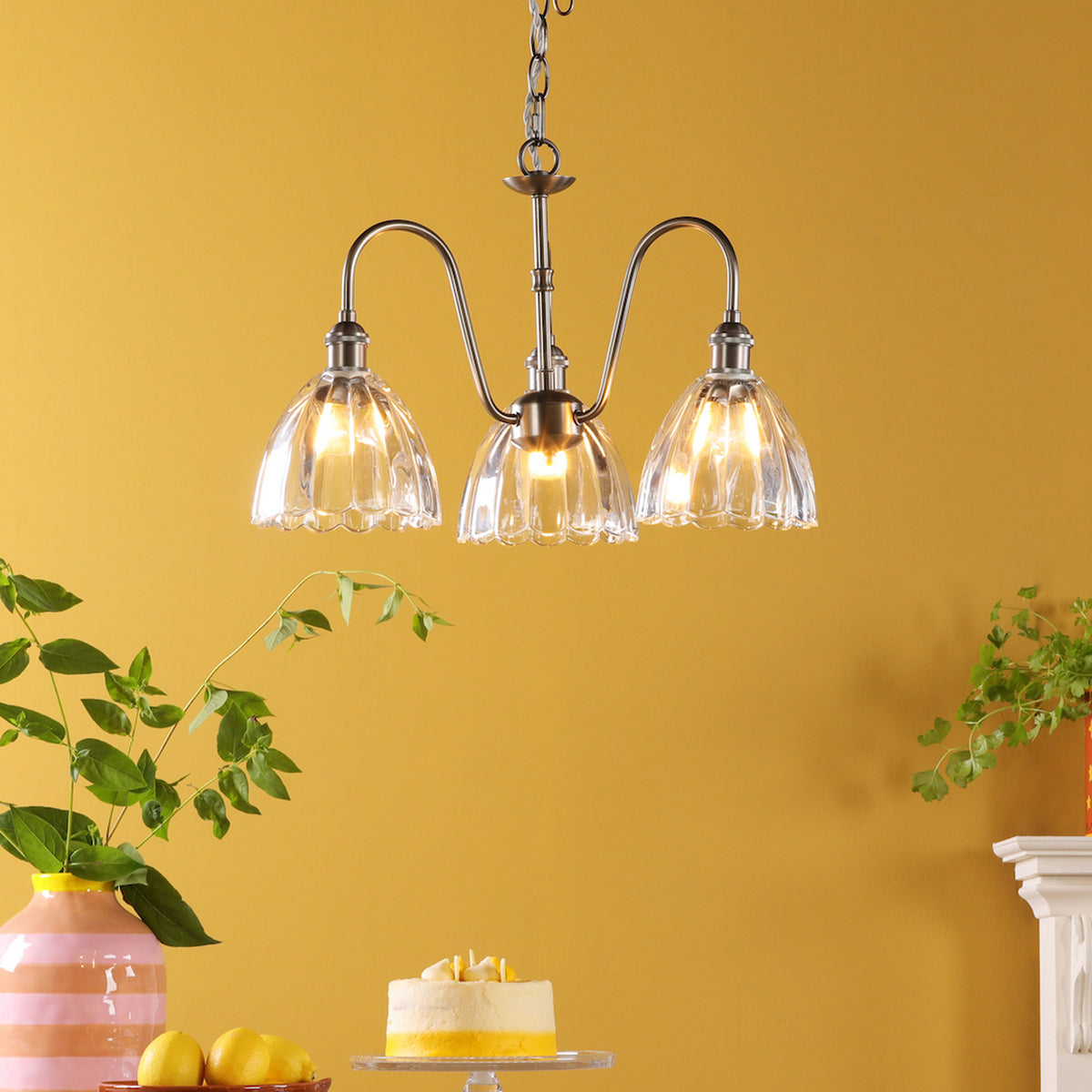 A three-light chandelier with clear, fluted glass shades hangs against a mustard yellow wall. Below, a lemon cake and lemons sit on a table.